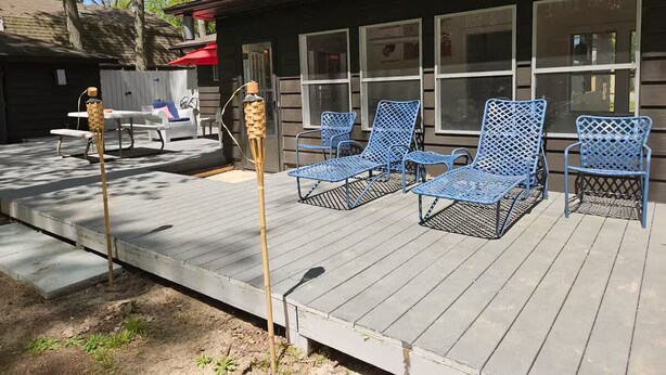En träveranda med blå metallstolar och ett bord, en bambustång och en byggnad med stora fönster i bakgrunden.