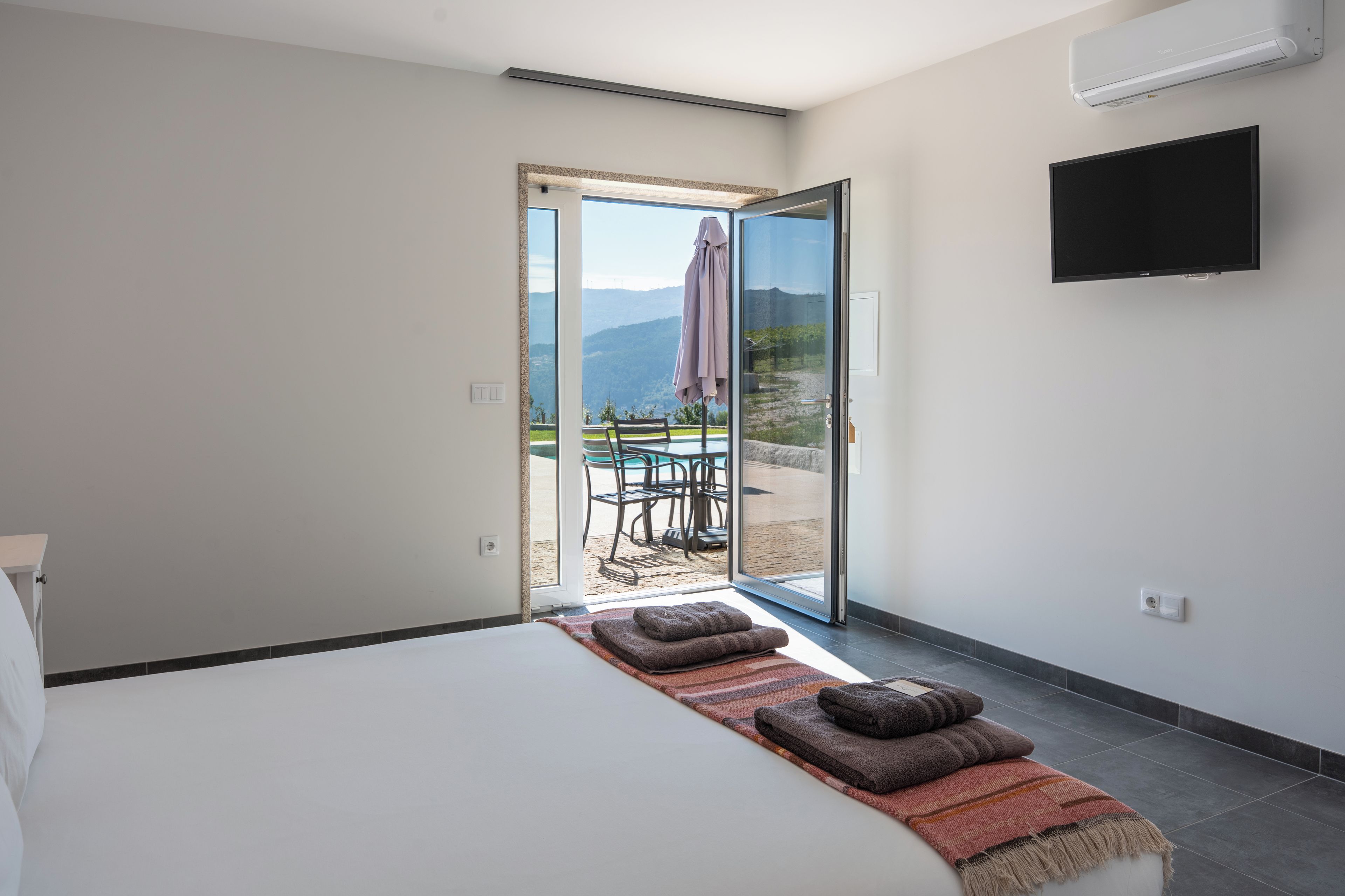 Un dormitorio con cama, televisión y puerta corrediza de vidrio que da a un área de estar exterior con mesa y sillas.