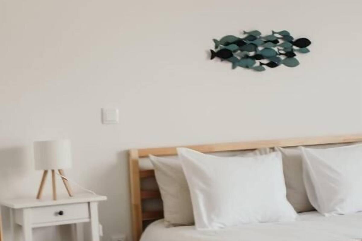 Ein Schlafzimmer mit Holzkopfteil, weißer Bettwäsche, einem kleinen Nachttisch mit Lampe und einer Wanddekoration aus grünen Blättern.