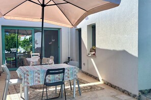 Une terrasse extérieure avec une table et des chaises sous un grand parasol, une porte-fenêtre coulissante et une petite fenêtre avec une étagère.