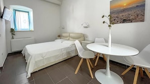 A bedroom with a bed, a bedside table, a chair, a small table, a window with curtains, a wall-mounted lamp, a painting of a sunset, and a potted plant. - B&B Dimora Orrù (Cabras)