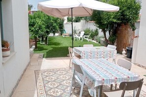 Un espace patio avec une table et des chaises sous une tonnelle, entouré d’une pelouse et d’un jardin bien entretenus.