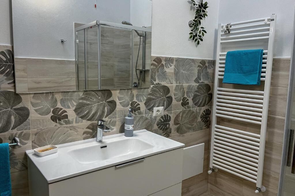 Un baño con un lavabo blanco, cabina de ducha de vidrio, toallero con una toalla azul y una pared con estampado de hojas.
