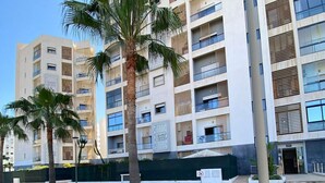 Een modern appartementencomplex met meerdere balkons, een aangelegde tuin met palmbomen en een verhard pad.