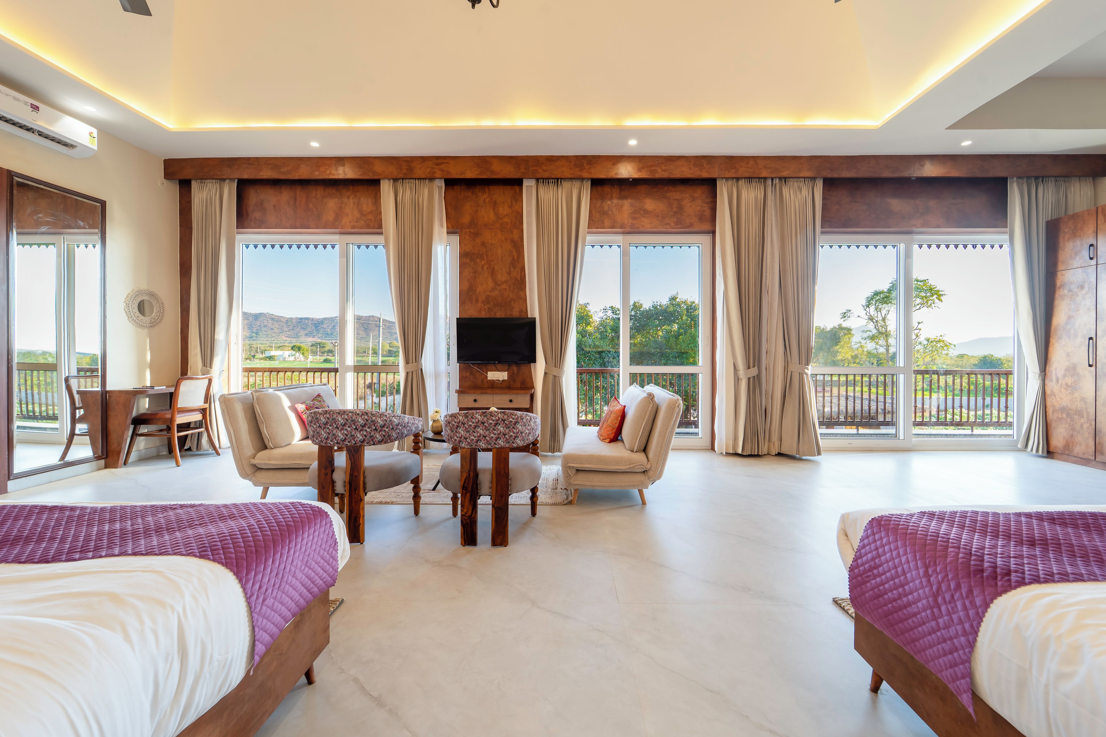 Amplia habitación de hotel con dos camas, zona de comedor, televisor y ventanas grandes con vistas al exterior.