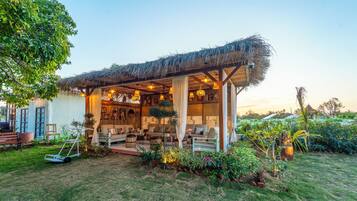 A covered outdoor seating area with a thatched roof, surrounded by greenery and a variety of seating options.