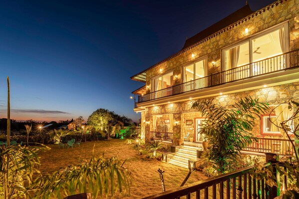 A stone building with a balcony, a staircase leading to the entrance, and a well-lit garden area.