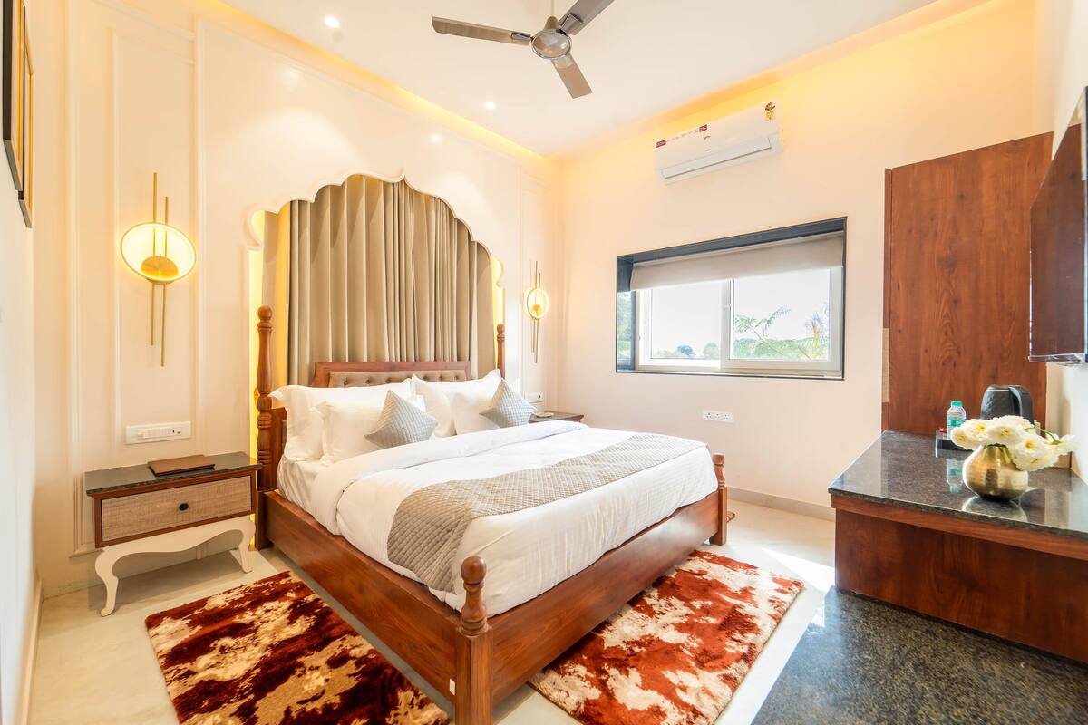 A bedroom with a wooden bed, a nightstand, a desk, and a window with a view of greenery.