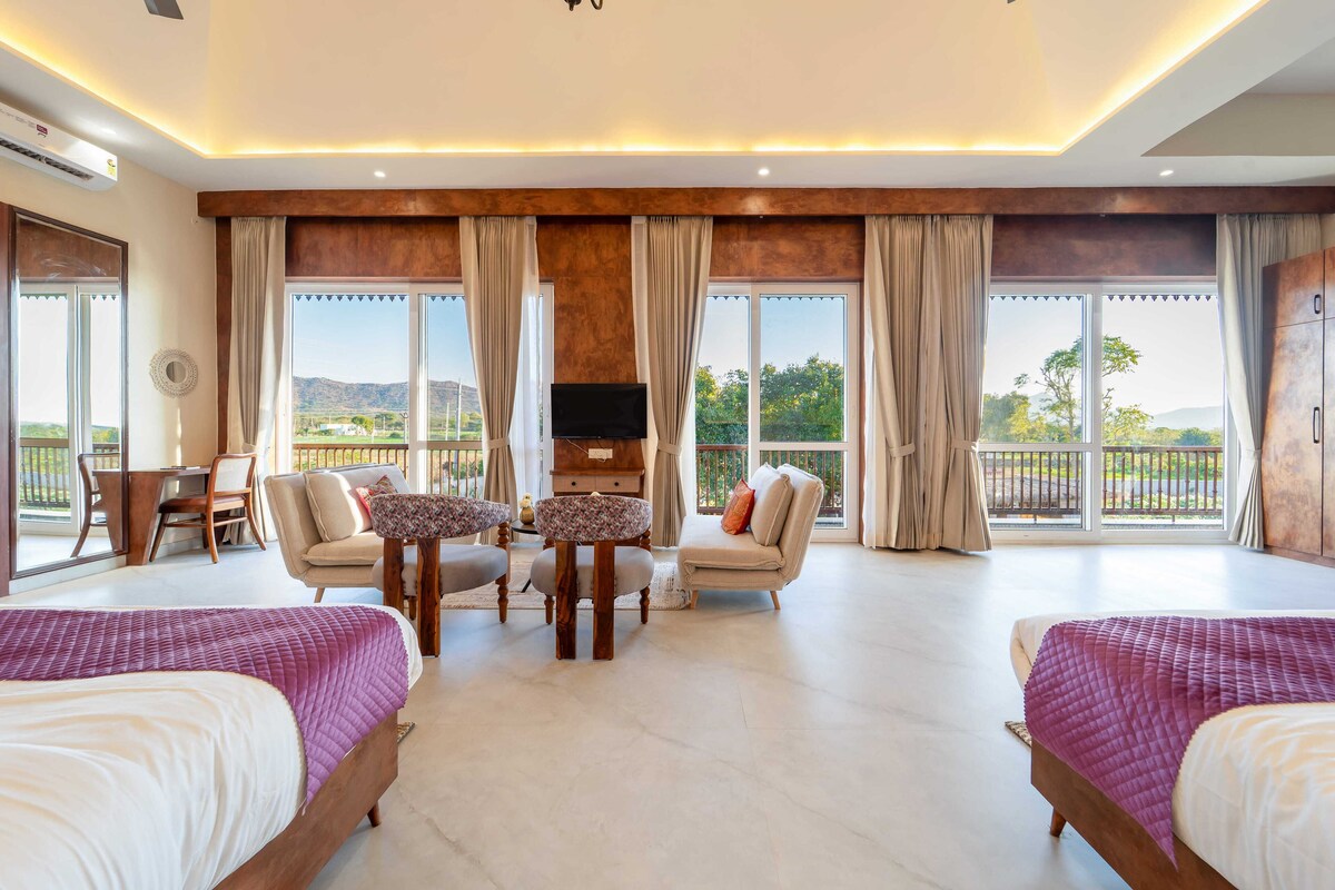 A spacious room with two beds, a TV, a dining table, and a balcony with a view of the mountains.