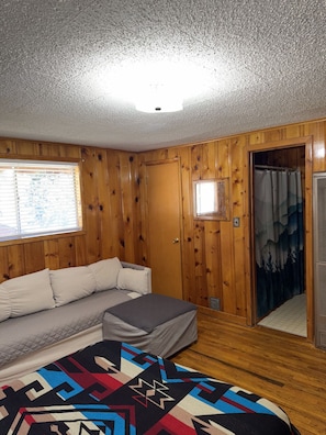 Una habitación con paredes de madera, un sofá, un tapete estampado y una puerta que da a otra habitación.