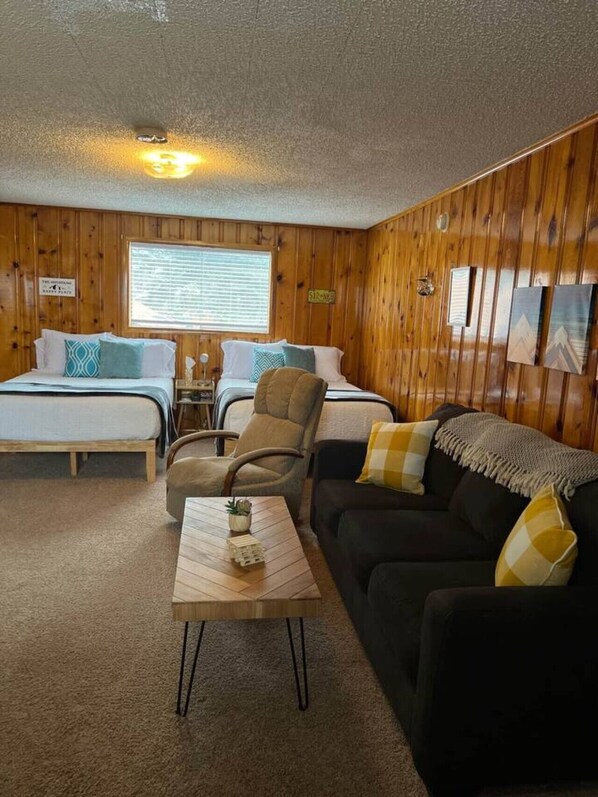 A room with wooden paneling, a bed with two pillows, a chair, a sofa, a small table, and a window with blinds. - Room 12 at Bellamy's (Red River)