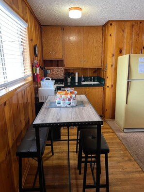A compact kitchen with wooden cabinets, a central island with stools, a refrigerator, and a window with blinds. - Room 12 at Bellamy's (Red River)