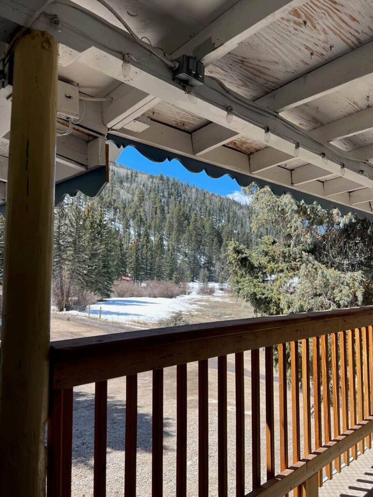A covered outdoor area with a wooden railing, a view of a forested mountain, and a clear sky.
