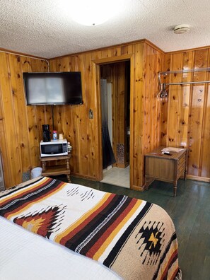 A room with wooden walls, a bed with a patterned blanket, a television mounted on the wall, a microwave, and a small wooden table.