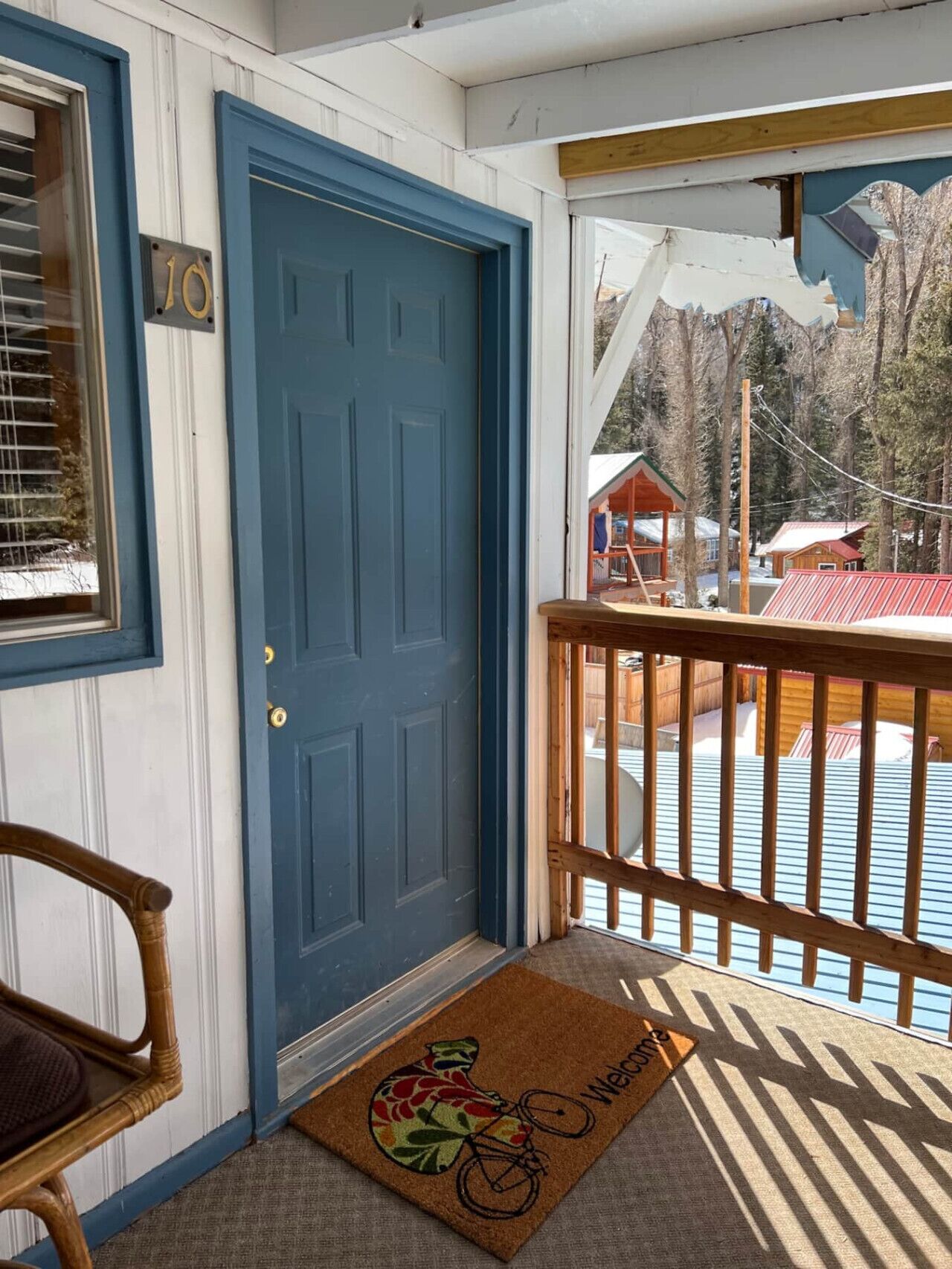 A blue door, a wooden chair, a doormat with a bicycle design, and a view of a cabin and trees.