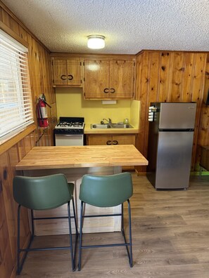 A compact kitchen with wooden cabinets, a gas stove, a sink, a refrigerator, and two green chairs. - Room 10 at Bellamy's (Red River)