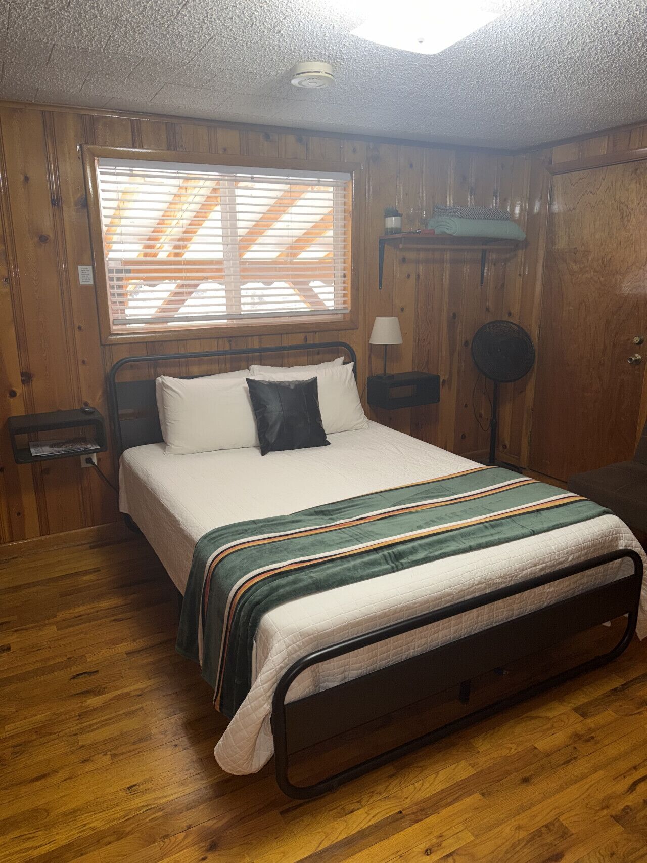 A wooden-paneled bedroom with a bed, a window with blinds, a bedside lamp, and a floor lamp.
