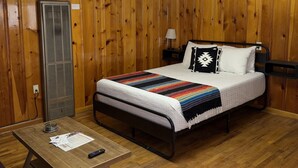 A room with wooden panel walls, a bed with a striped blanket, a bedside table with papers, and a heater. - Room 5 at Bellamy's Haus (Red River)