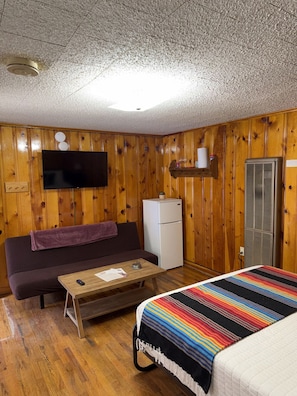 Una habitación con paneles de madera, un televisor de pantalla plana montado en la pared, un refrigerador pequeño, un sofá con una manta, una mesita de centro y una cama con una colcha a rayas.