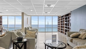 A spacious room with a large window overlooking the sea, a comfortable seating area, and a bookshelf. - Tidewater Horizon by Avantstay Beachfront Condo + Indoor Pool + Spa Amenities + Movie Theater + Near Pier Park (Panama City Beach)
