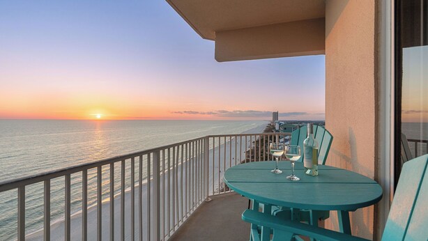 A balcony with a view of the sunset over the ocean, a table with two glasses of wine, and a bottle of wine. - Tidewater Horizon by Avantstay Beachfront Condo + Indoor Pool + Spa Amenities + Movie Theater + Near Pier Park (Panama City Beach)