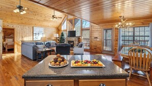 A cozy cabin interior with a fireplace, a dining area with fruit, a sofa, and a large window. - Grand Getaway Cabin by Avantstay A-frame Retreat + Hot Tub + Pool Access, Game Room Mins to Dollywood (Sevierville)