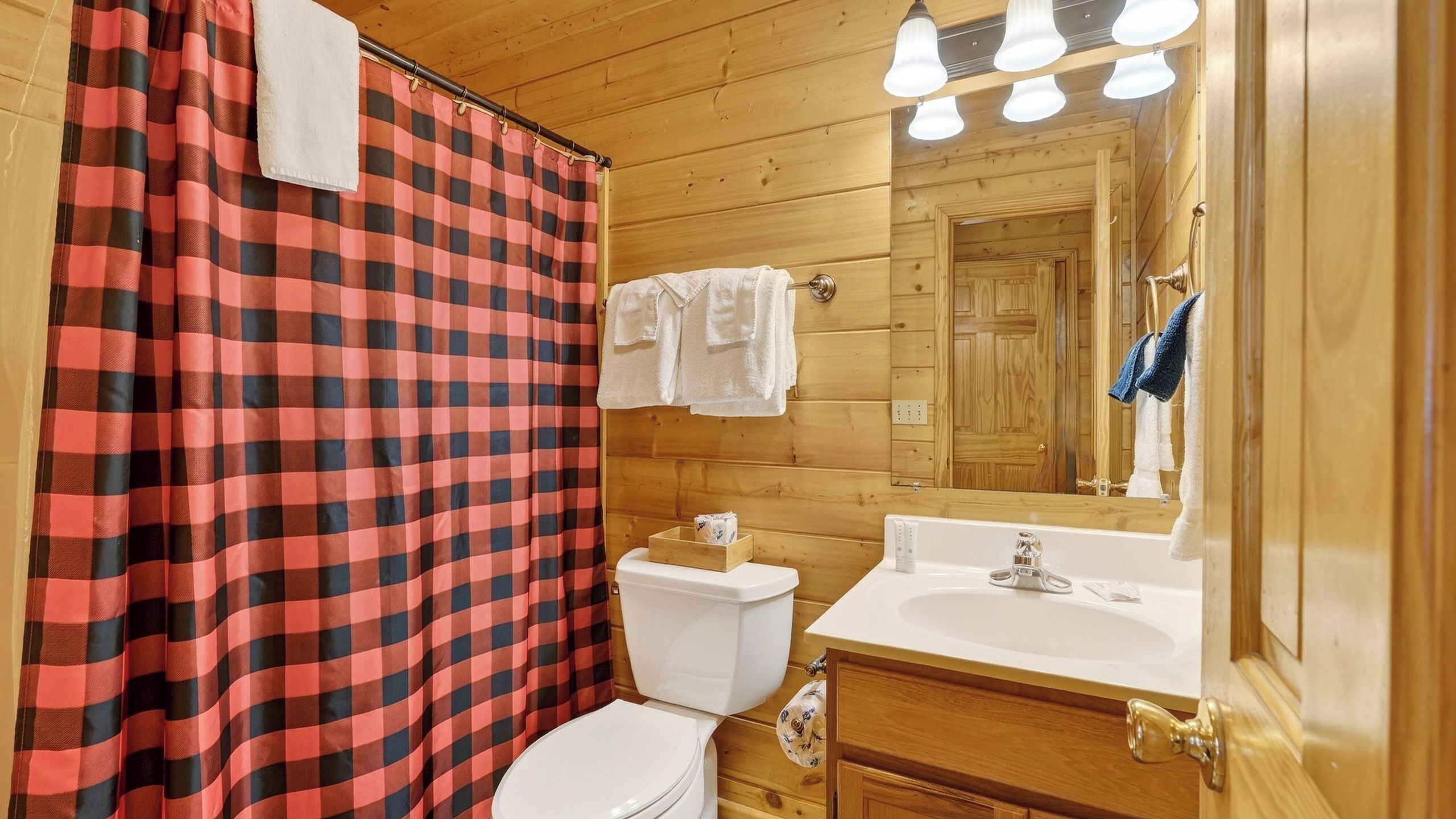 A bathroom with a checkered shower curtain, a toilet, a sink with a mirror, and wooden walls.