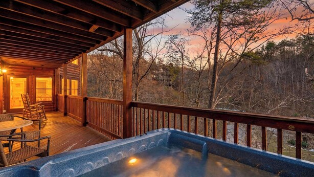 A wooden deck with a hot tub, chairs, and a view of trees and a sunset. - Grand Getaway Cabin by Avantstay A-frame Retreat + Hot Tub + Pool Access, Game Room Mins to Dollywood (Sevierville)