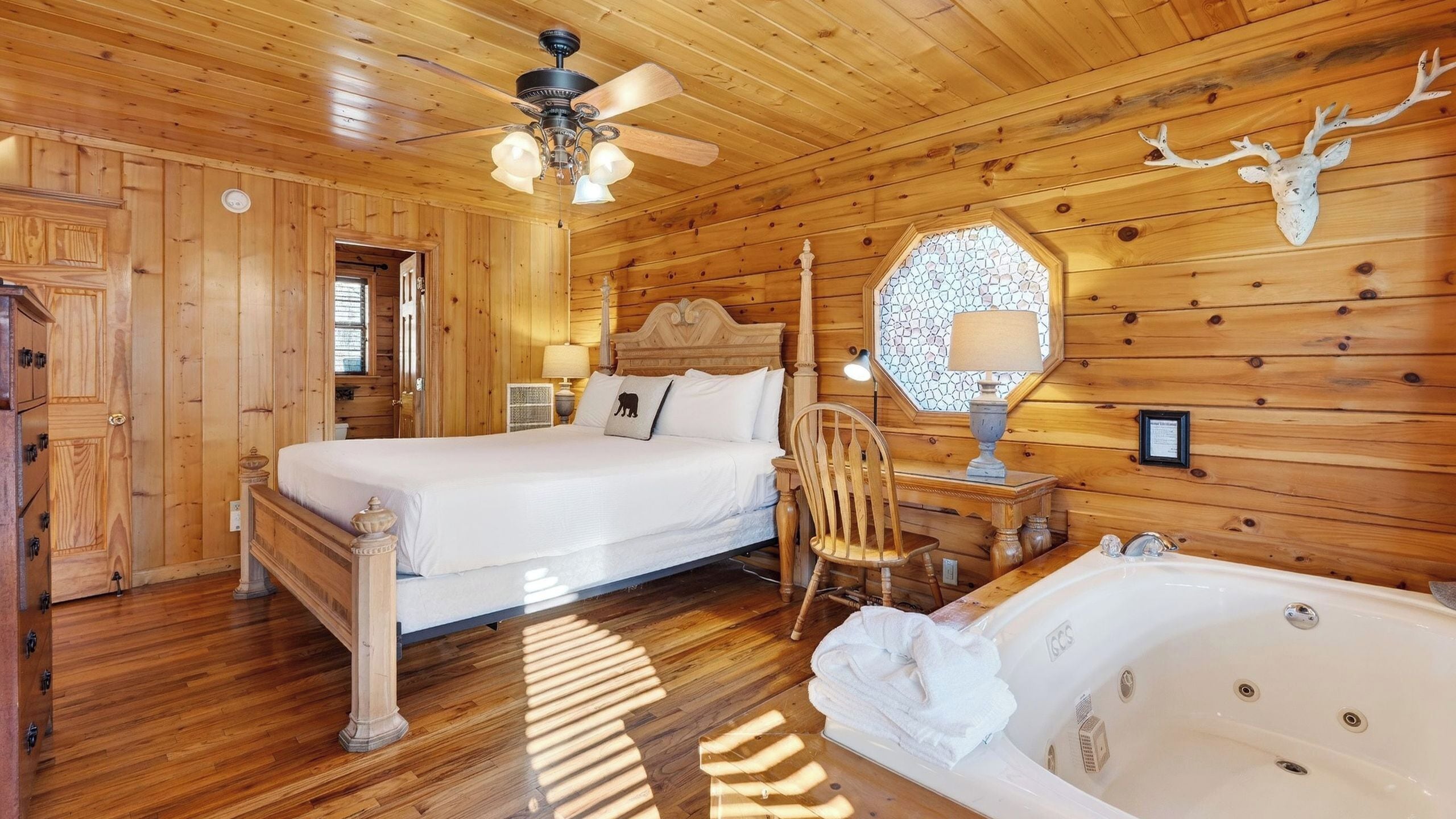 Een slaapkamer in een houten hutje met een bed, een stoel, een nachtkastje, een lamp, een decoratie van een hertenkop en een bad.
