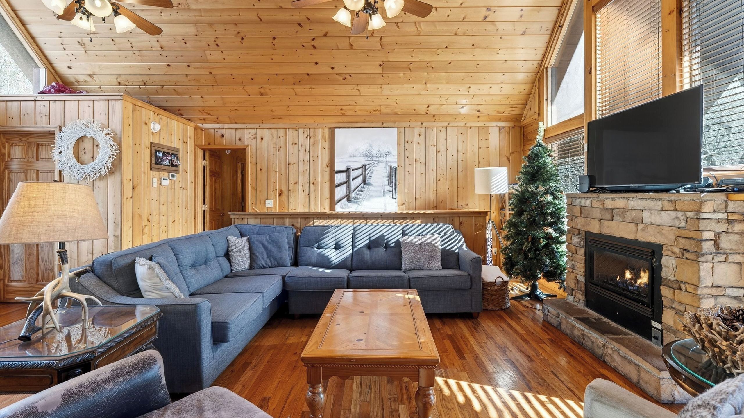 A cozy cabin interior with a wooden ceiling, a sectional sofa, a fireplace, and a television.