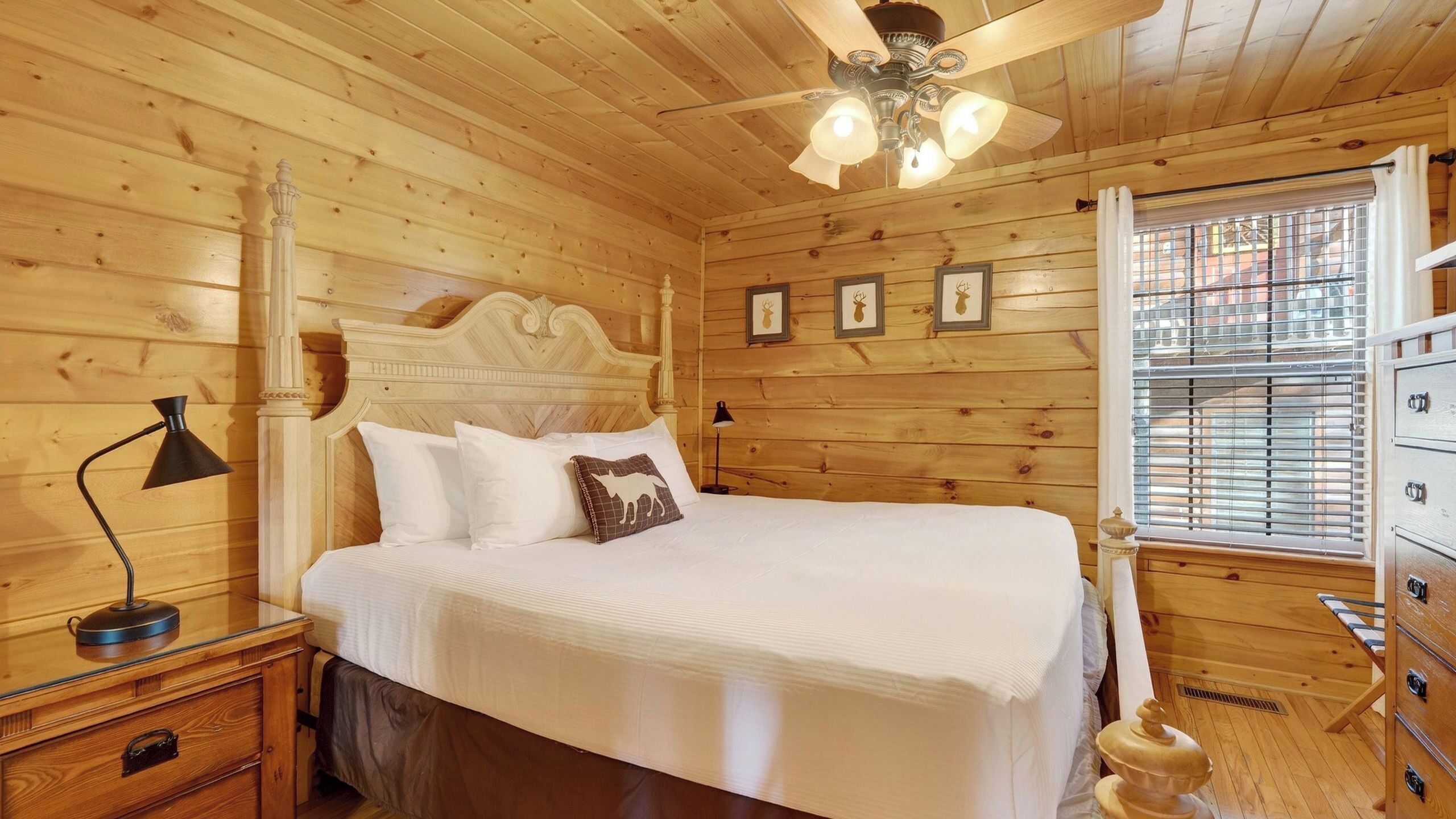 Un dormitorio de cabaña de madera con una cama, una mesita de noche, una lámpara, un ventilador de techo y una ventana con persianas.