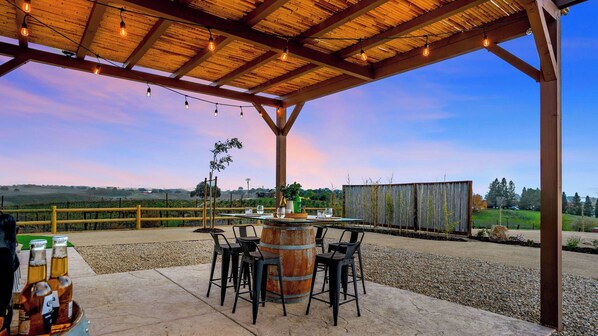 An outdoor patio with a wooden pergola, string lights, a rustic barrel table, and chairs. - Sip and Stay at Vahali Vineyards by Avantstay Modern Wine Country Duo + Outdoor Dining + Vineyard Views (Paso Robles)