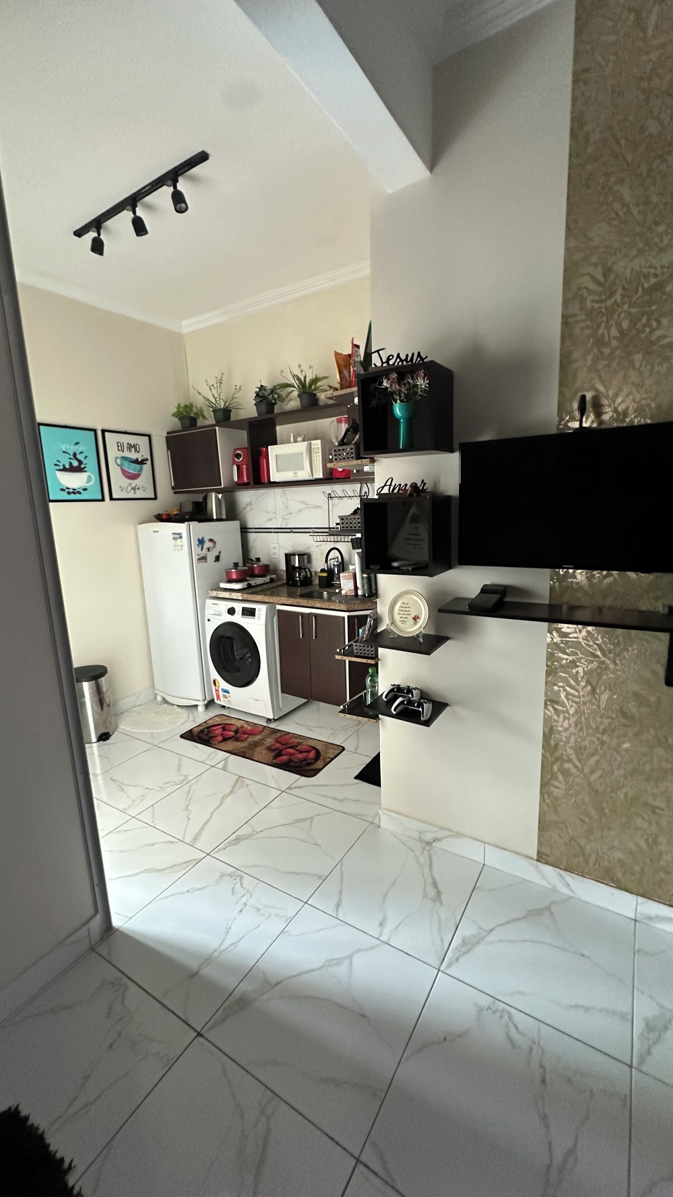 Cocina funcional con lavadora, microondas y refrigerador pequeño. Hay plantas en macetas y una estantería con objetos decorativos.