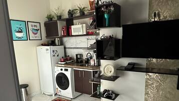 Cocina funcional con lavadora, microondas y refrigerador pequeño. Hay plantas en macetas y una estantería con objetos decorativos.