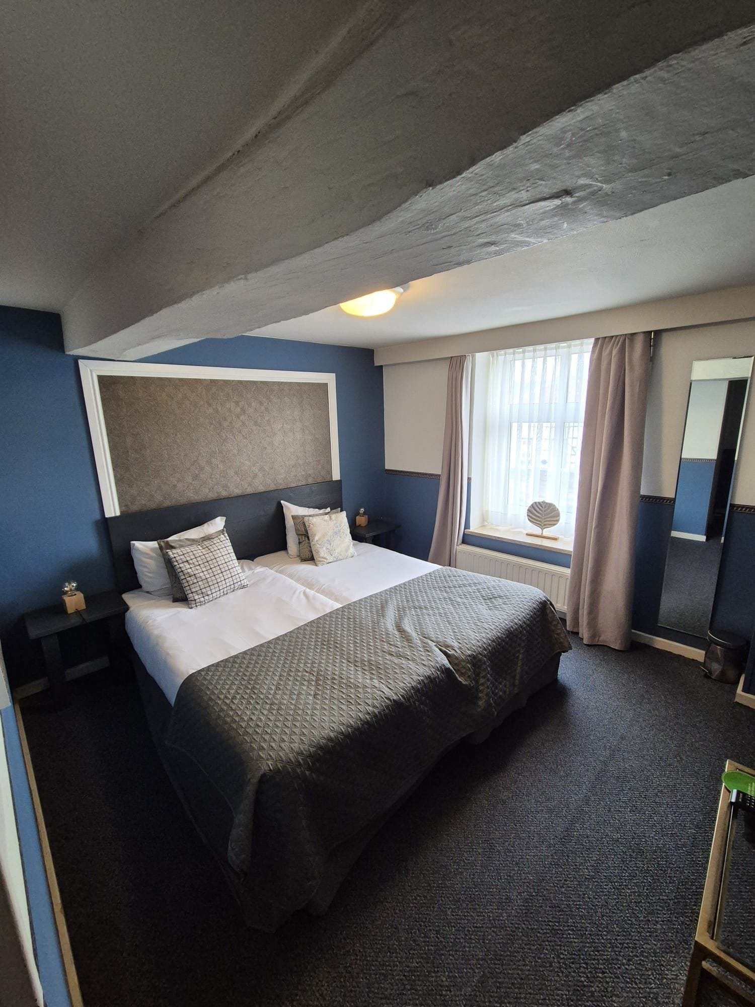 Une chambre d’hôtel avec un grand lit, une table de chevet, une commode, une fenêtre avec des rideaux et un miroir.