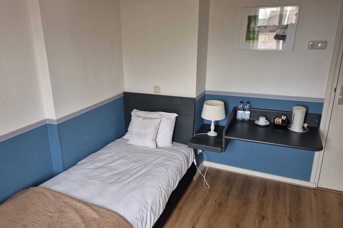 Una habitación pequeña con cama individual, mesita de noche, lámpara y espejo.