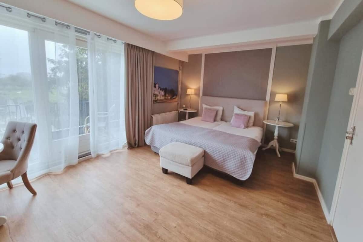 Un dormitorio con una cama grande, dos mesitas de noche, una silla y vistas al exterior a través de cortinas translúcidas.