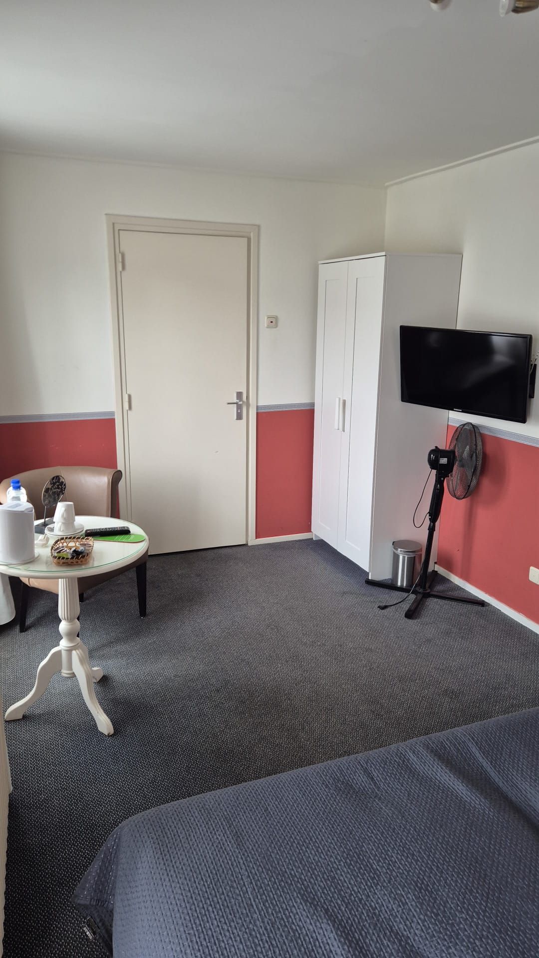 A hotel room with a bed, a table with a basket, a chair, a TV mounted on the wall, and a standing fan.