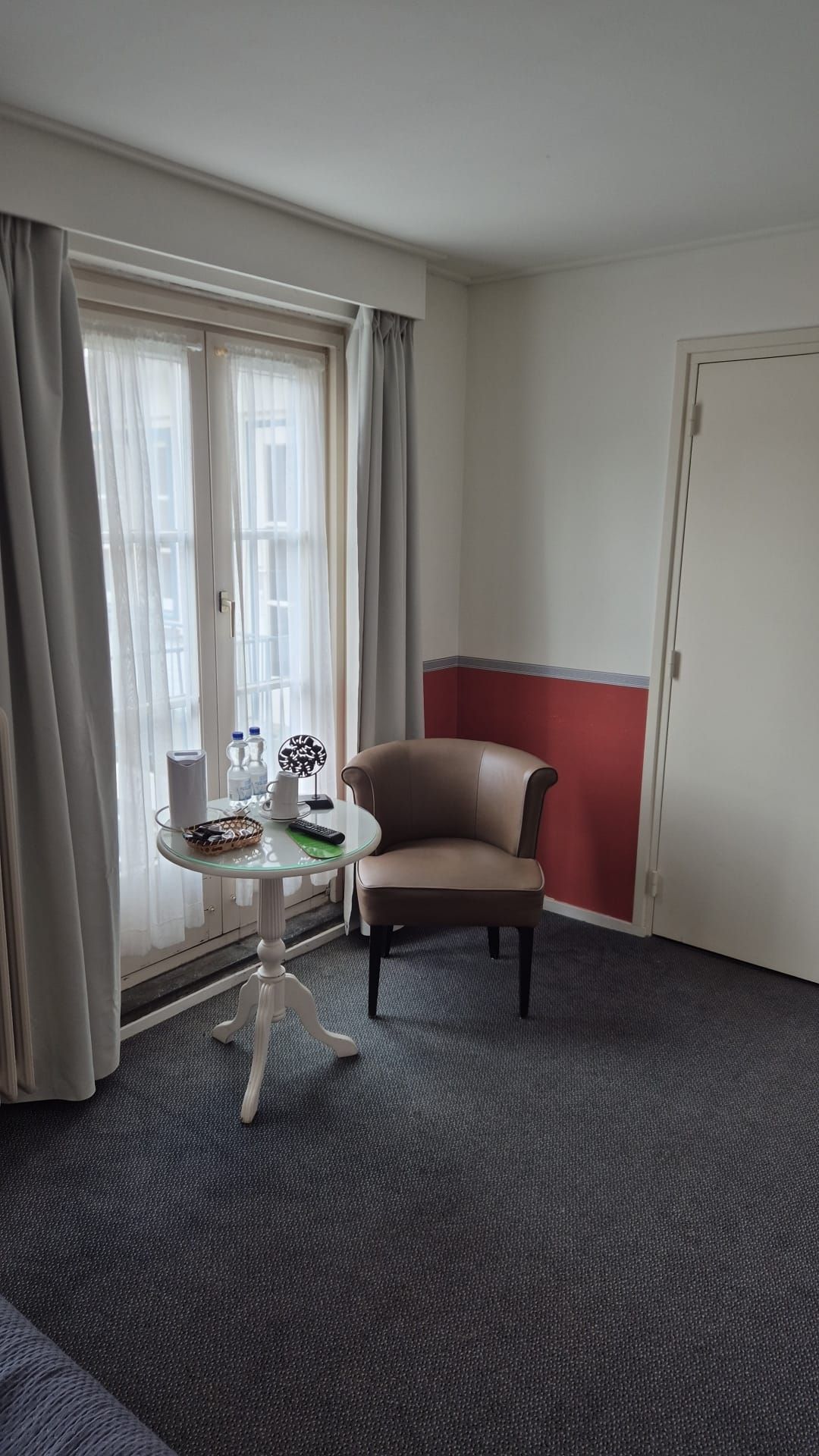 A hotel room with a single chair, a small table with a remote control, and a window with curtains.