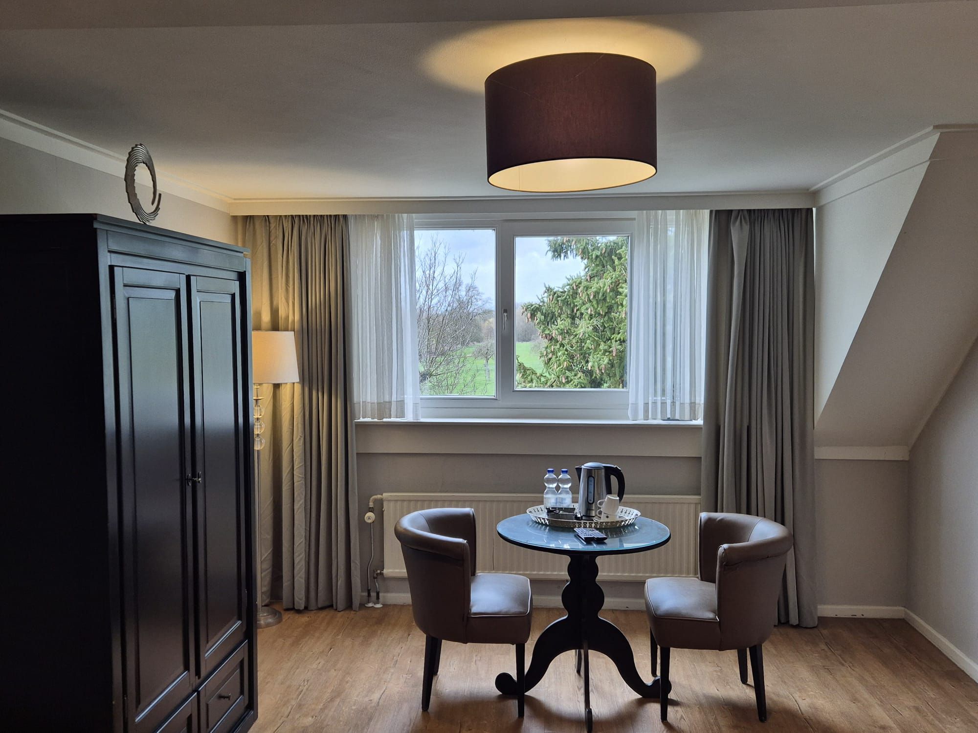 Ein Zimmer mit einem Tisch, zwei Stühlen, einem Schrank, einem Fenster mit Vorhängen und einer Deckenleuchte.