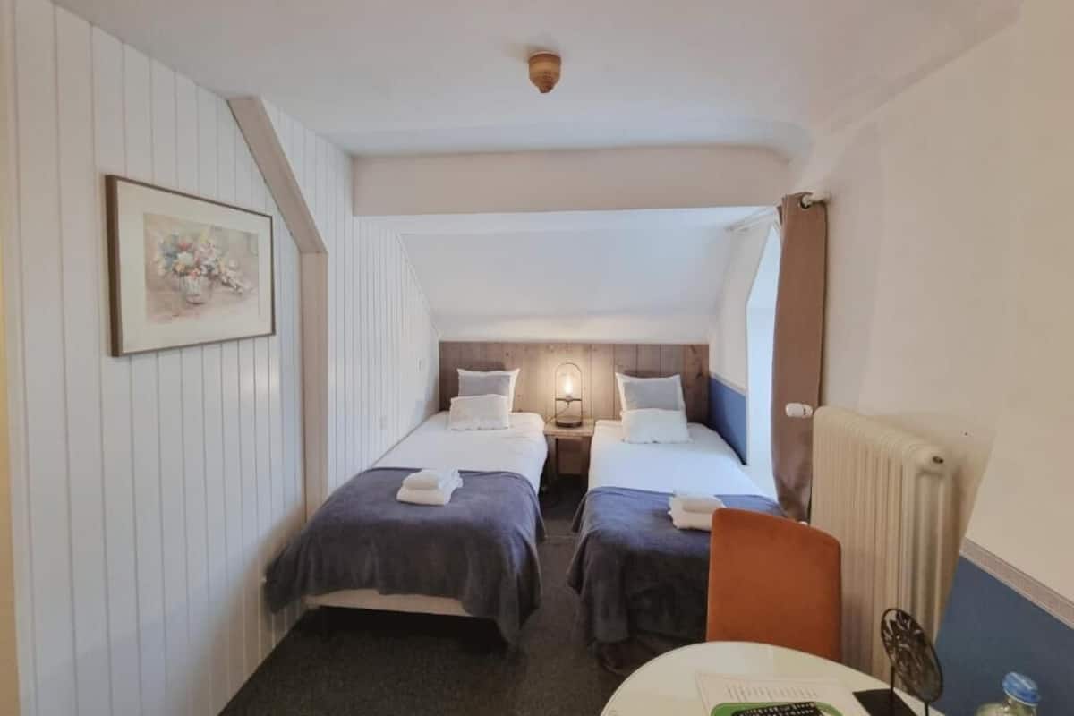Una habitación de hotel con dos camas individuales, una mesita con una silla, un cuadro en la pared y un radiador.