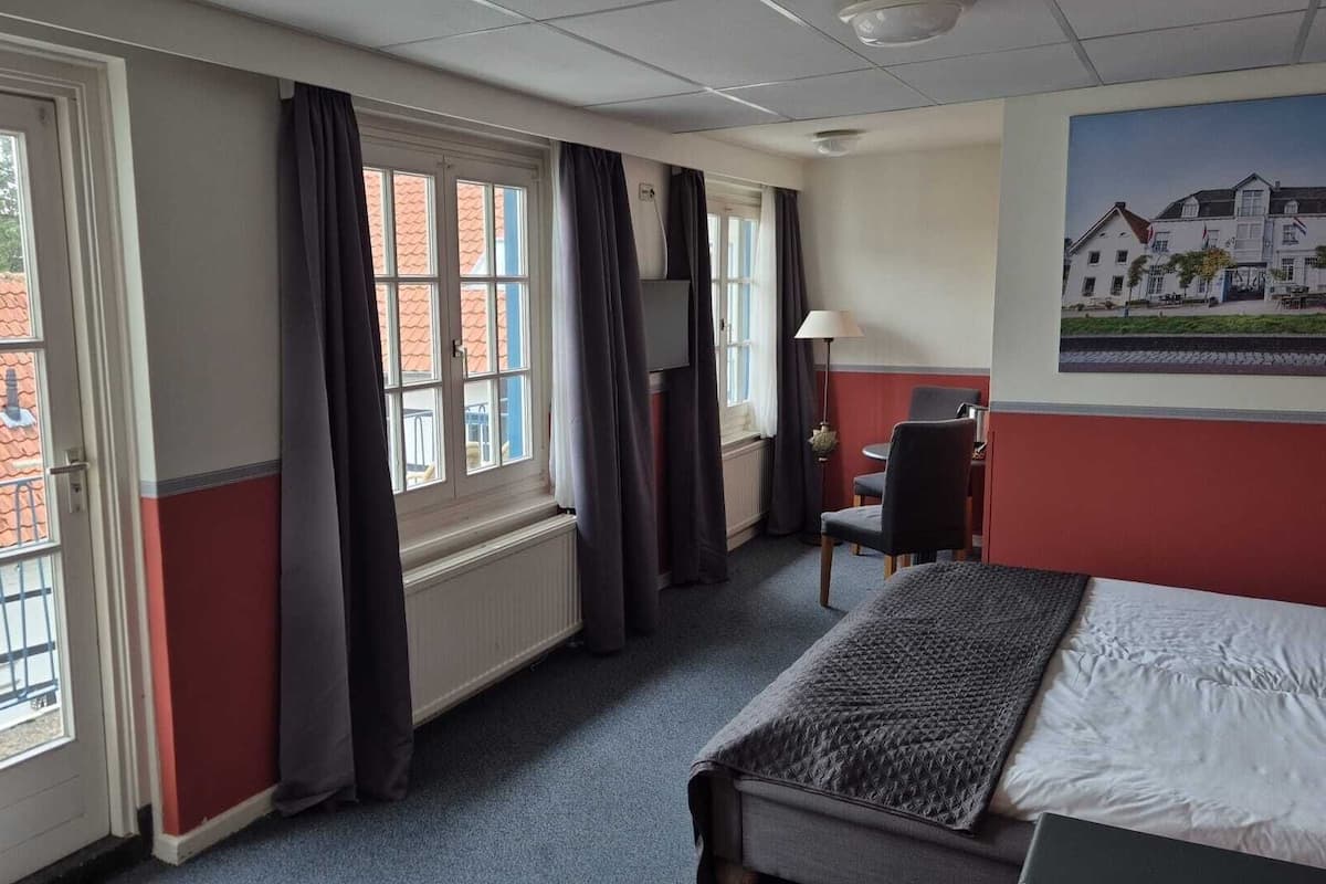 Habitación de hotel con cama, escritorio, silla, ventana con cortinas y un cuadro en la pared.
