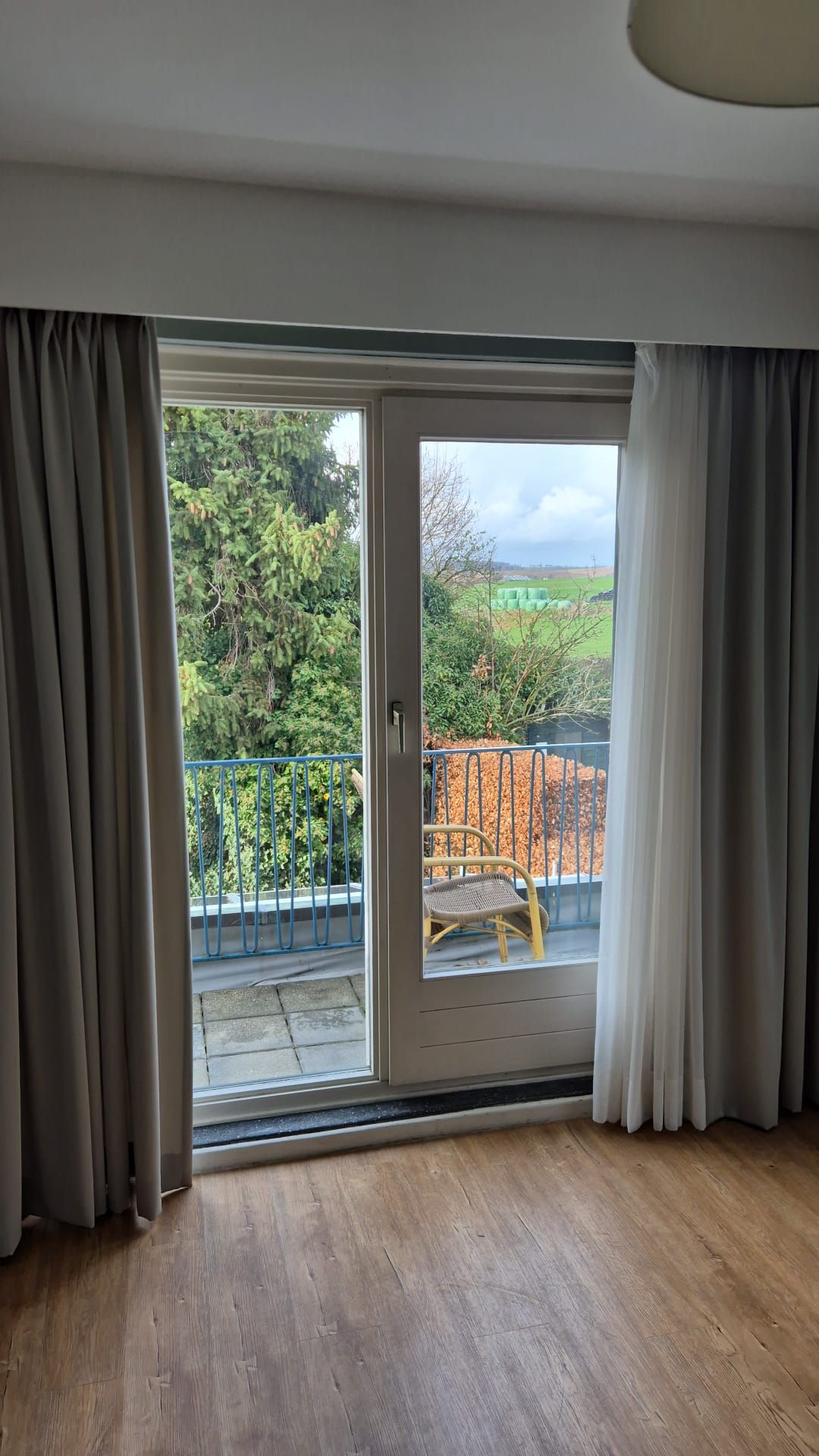 A room with a wooden floor, a sliding glass door leading to a balcony with a chair and a metal railing.