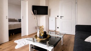 Un saló modern amb una taula de centre de vidre, un sofà negre, un llit, un televisor muntat a la paret i una porta blanca.