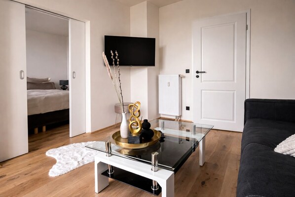 Ein modernes Wohnzimmer mit einem Glastisch, einem schwarzen Sofa, einem Bett, einem an der Wand montierten Fernseher und einer weißen Tür.