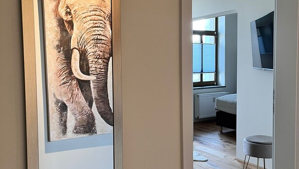 Un passadís amb una obra d'art emmarcada d'un filó, que condueix a una habitació amb un llit, una televisió i una finestra.