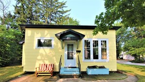 A small, single-story yellow house with a white door and windows, a front porch with a bench, and a small garden area. - New Listing: perfect location, Sunshine Cottage,  a cute little gem in Lakeside! (Muskegon)