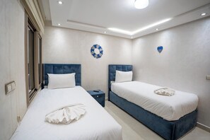 A hotel room with two beds, each with a blue headboard, white bedding, and a towel on the beds. There is a small nightstand between the beds, a wall decoration, and a ceiling light. - Nathalie (Marrakech)
