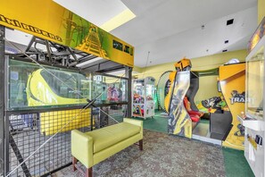 Una habitación con un sofá amarillo, un separador de vidrio y máquinas de videojuegos.