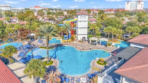 A large outdoor swimming pool surrounded by palm trees and lounge chairs, with a water slide and a building with a tower in the background. - Power Up Palace | Disney, River & Slides (Kissimmee)